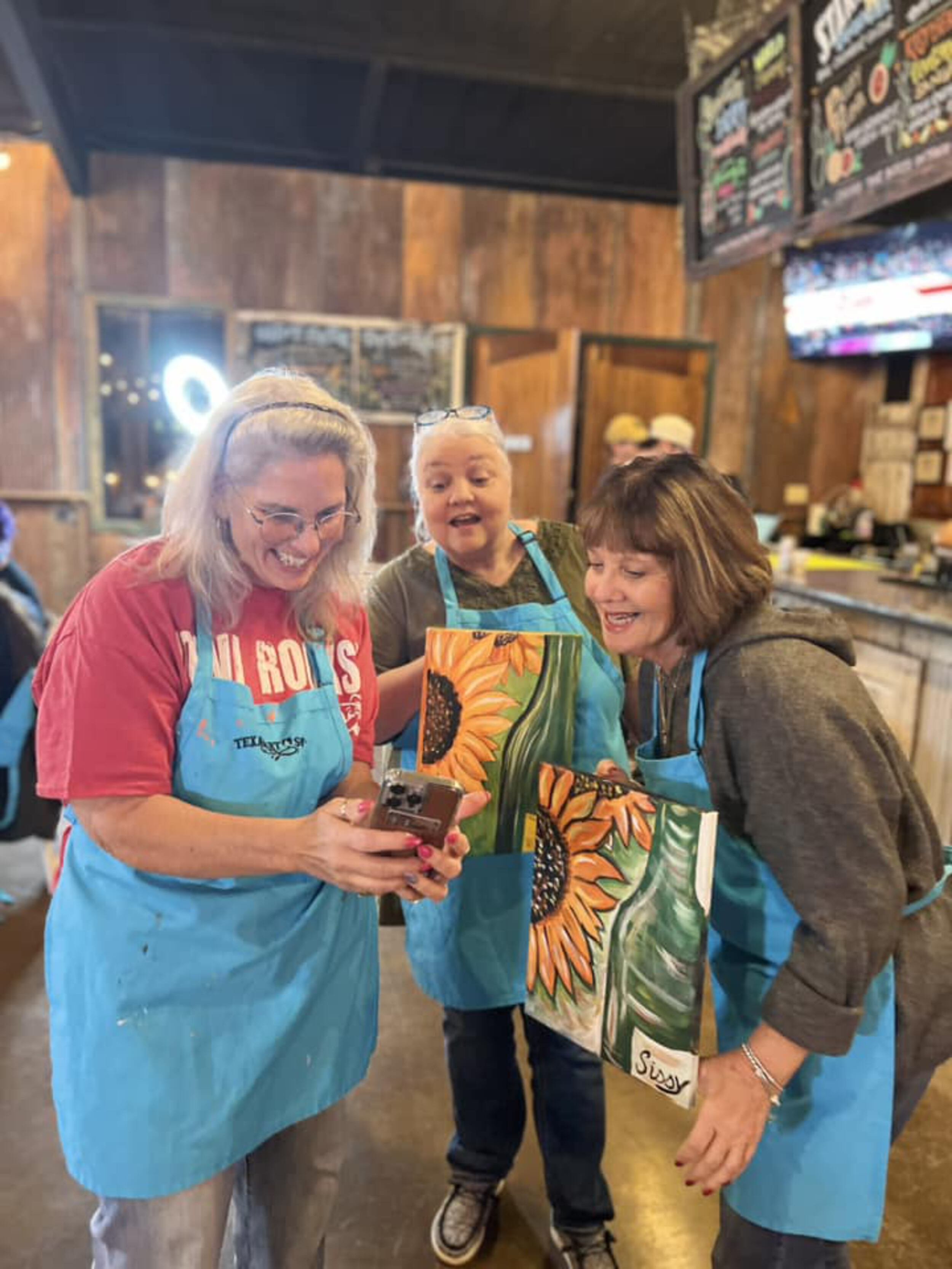 Three women in a rustic indoor setting, wearing blue aprons, appearing joyful and looking at a phone, holding tote bags with sunflower designs.