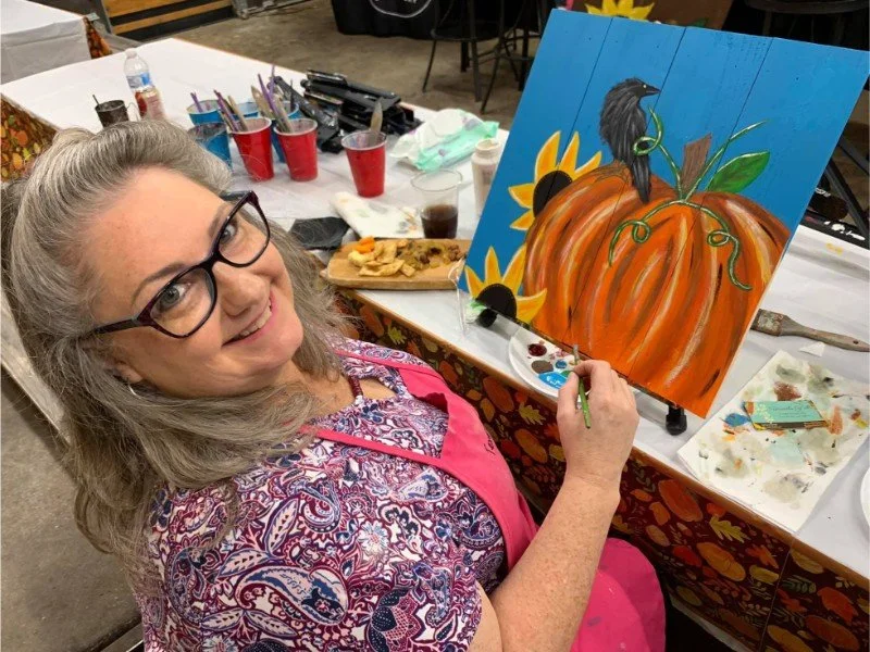 Woman with glasses painting a pumpkin on a canvas at an art party.