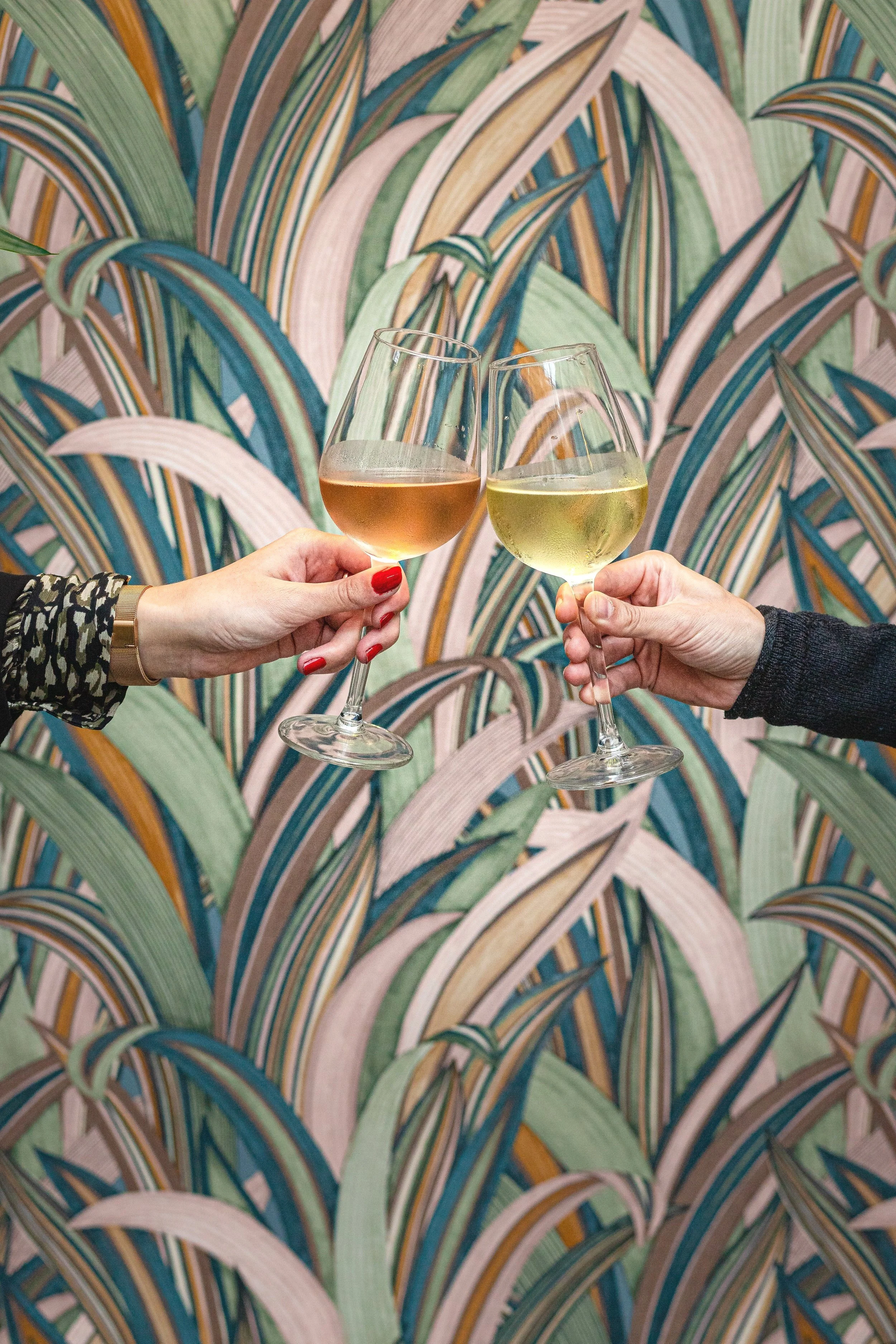 Two people clinking glasses of wine against a colorful leafy patterned background.