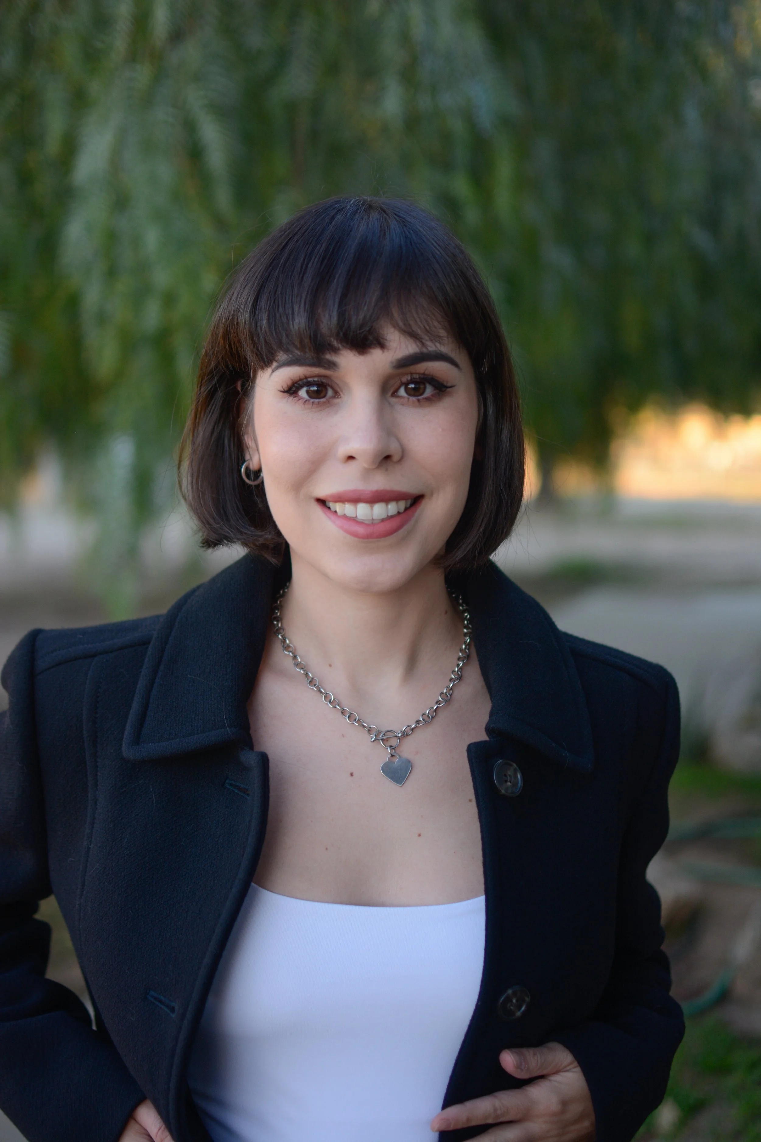 A woman with short dark hair and light skin, smiling outdoors with trees in the background. She is wearing a black jacket, a white top, hoop earrings, and a silver necklace with a heart pendant.