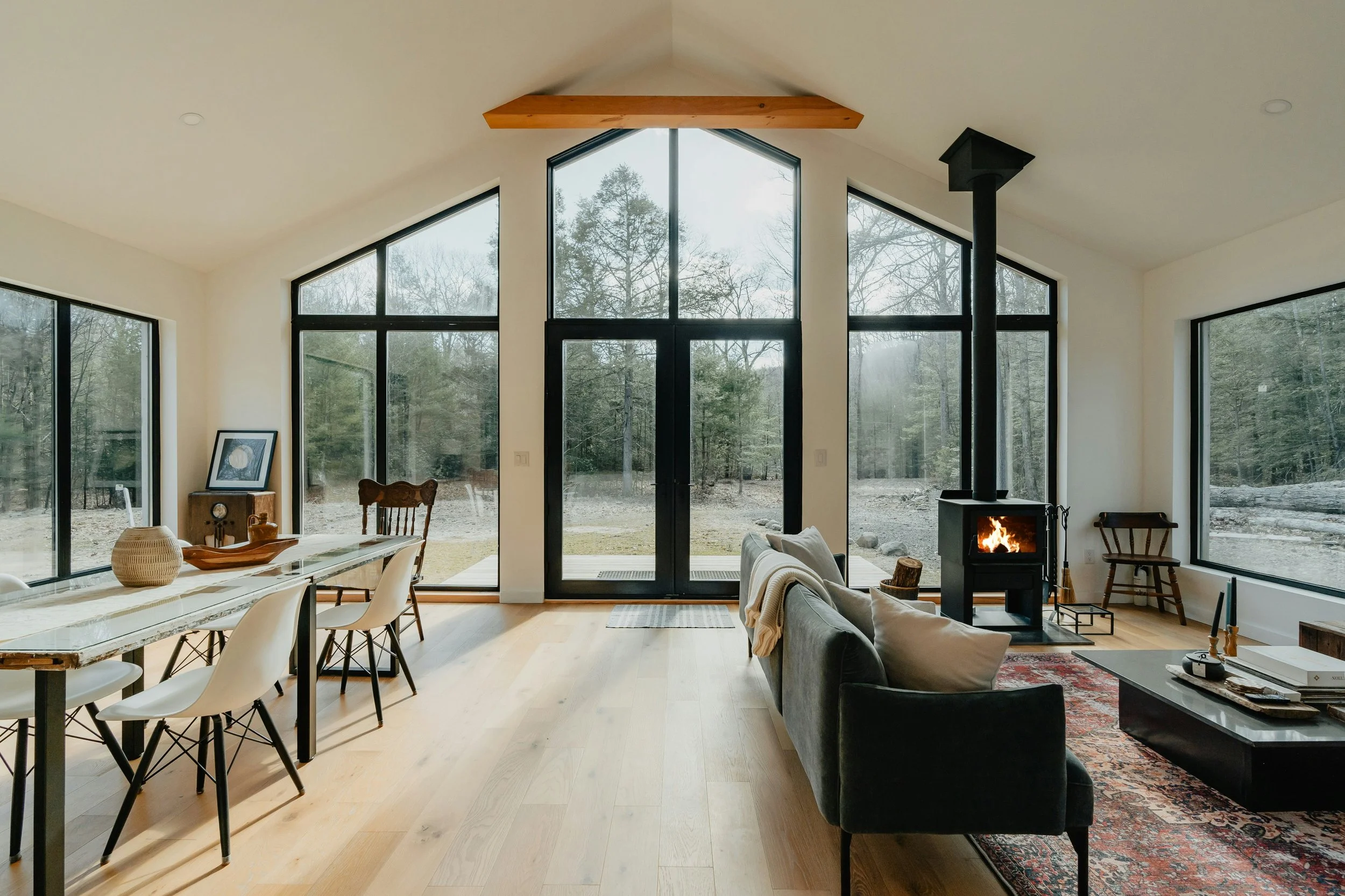Living room with large windows and a glass door, wood floors, a black wood stove with a fire, a gray sofa, a coffee table, and a dining table with chairs, with a view of a wooded outdoor area.