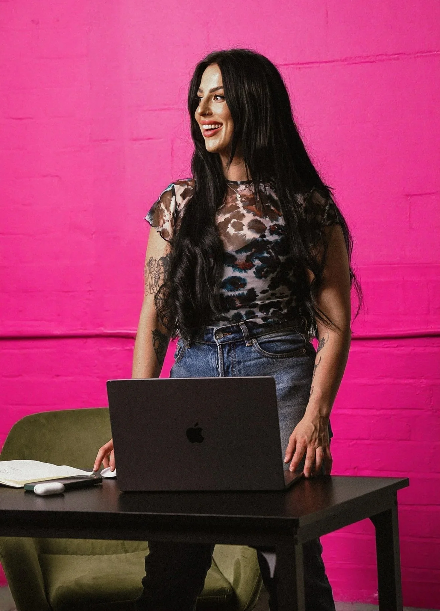 A woman with long black hair, a leopard print top, and tattoos on her arms, standing behind a black table with an open MacBook, in front of a bright pink wall.