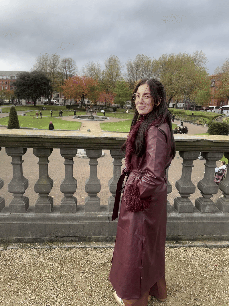A woman with long dark hair and glasses wearing a maroon trench coat stands on a stone balcony overlooking a park with trees and a fountain on a cloudy day.