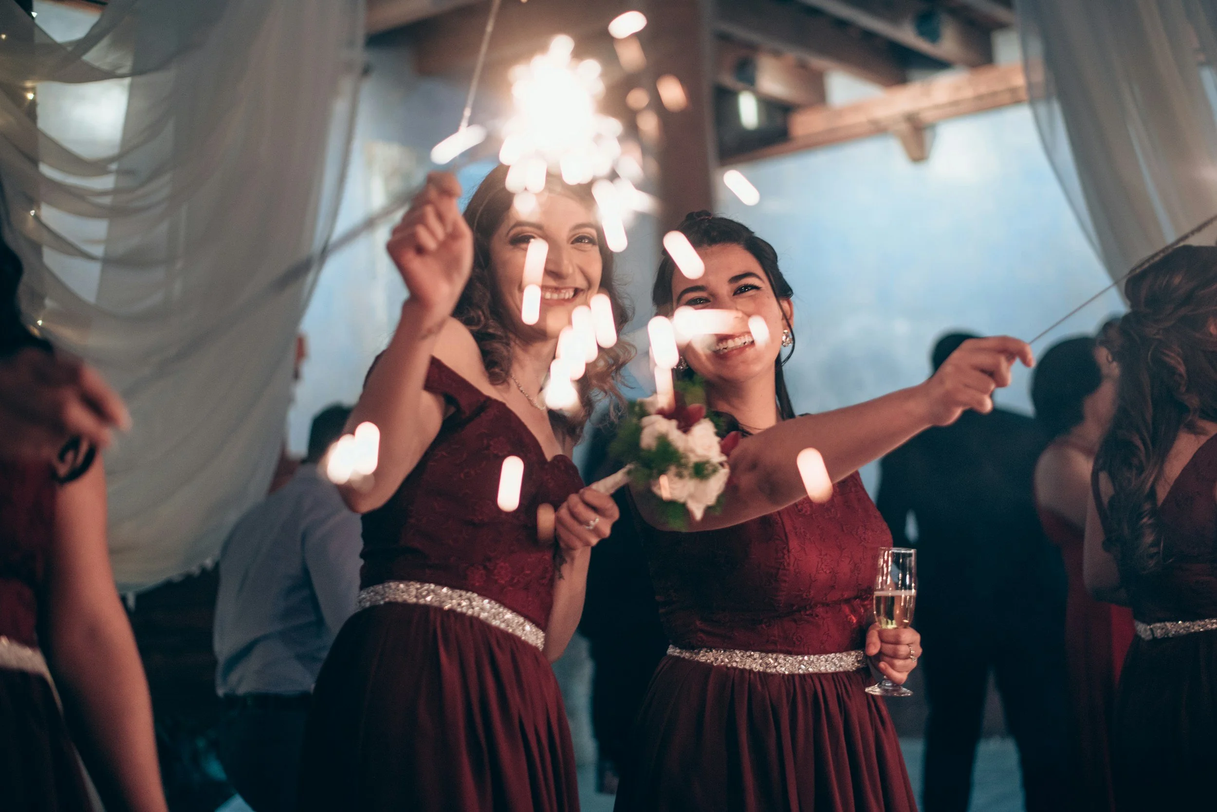 Two women in red dresses celebrating at a party, holding sparklers and a glass of champagne, smiling and enjoying themselves.