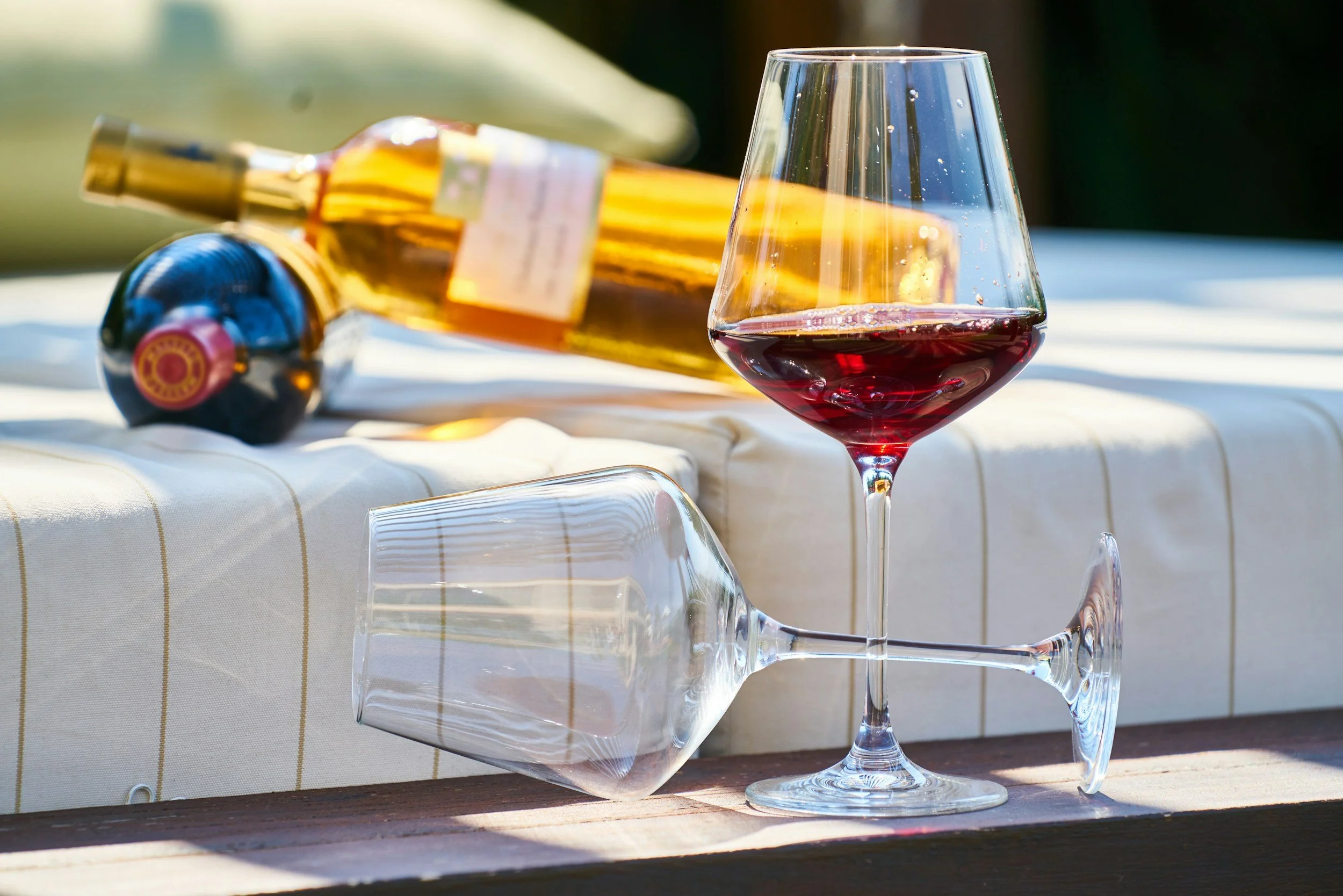 A glass of red wine, a wine bottle, and an empty wine glass are set on a table outdoors, with a blurred background of greenery.