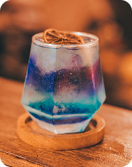 Colorful layered cocktail in a short glass with ice and a lemon slice, on a wooden coaster.
