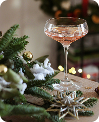 A pink cocktail in a coupe glass on a wooden surface with Christmas decorations, including a pine branch, ornaments, and a star-shaped decoration.