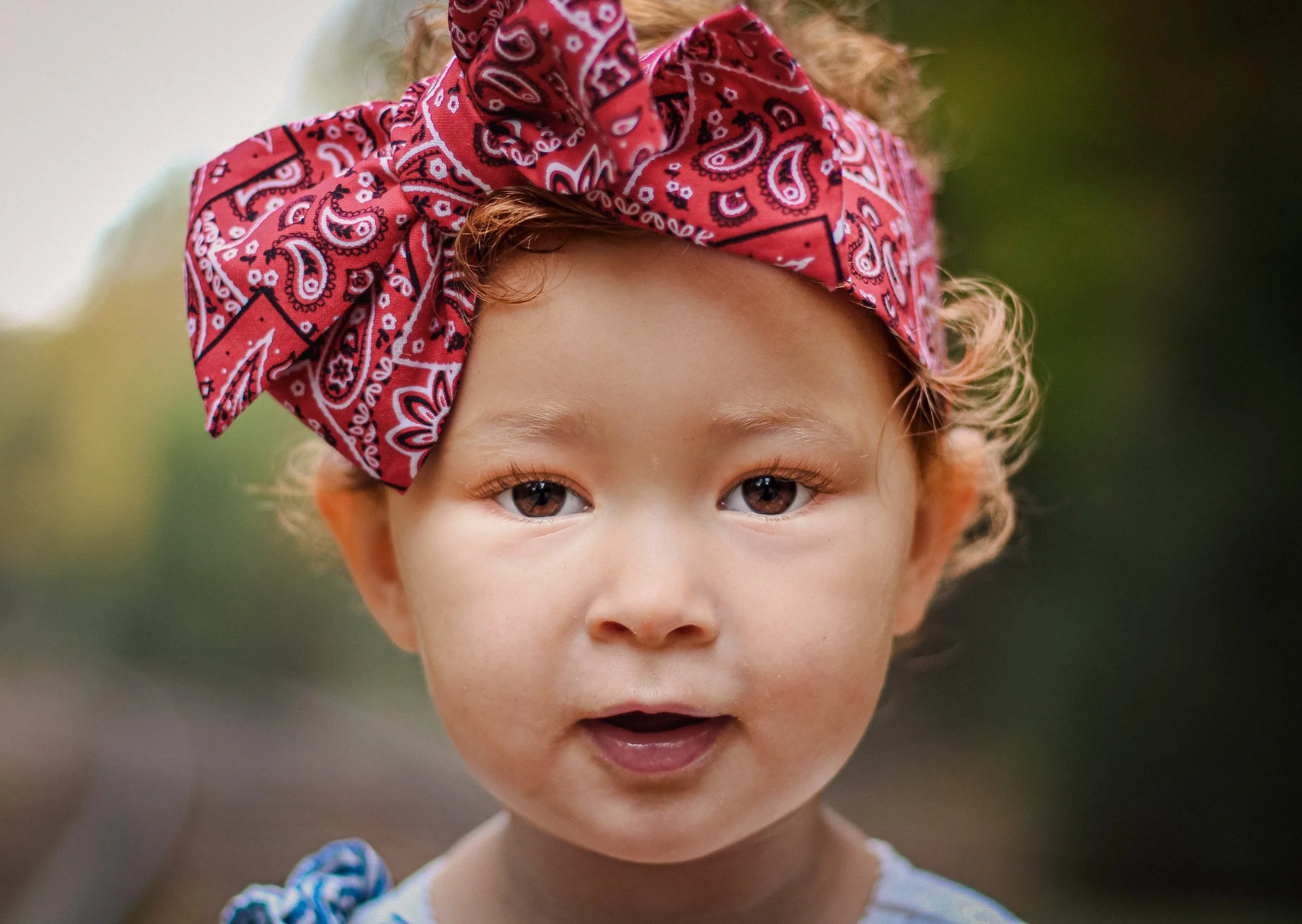Close-up of a young child with light skin, brown eyes, and curly hair wearing a red bandana with white paisley patterns tied around their head, outdoors with blurred nature background.
