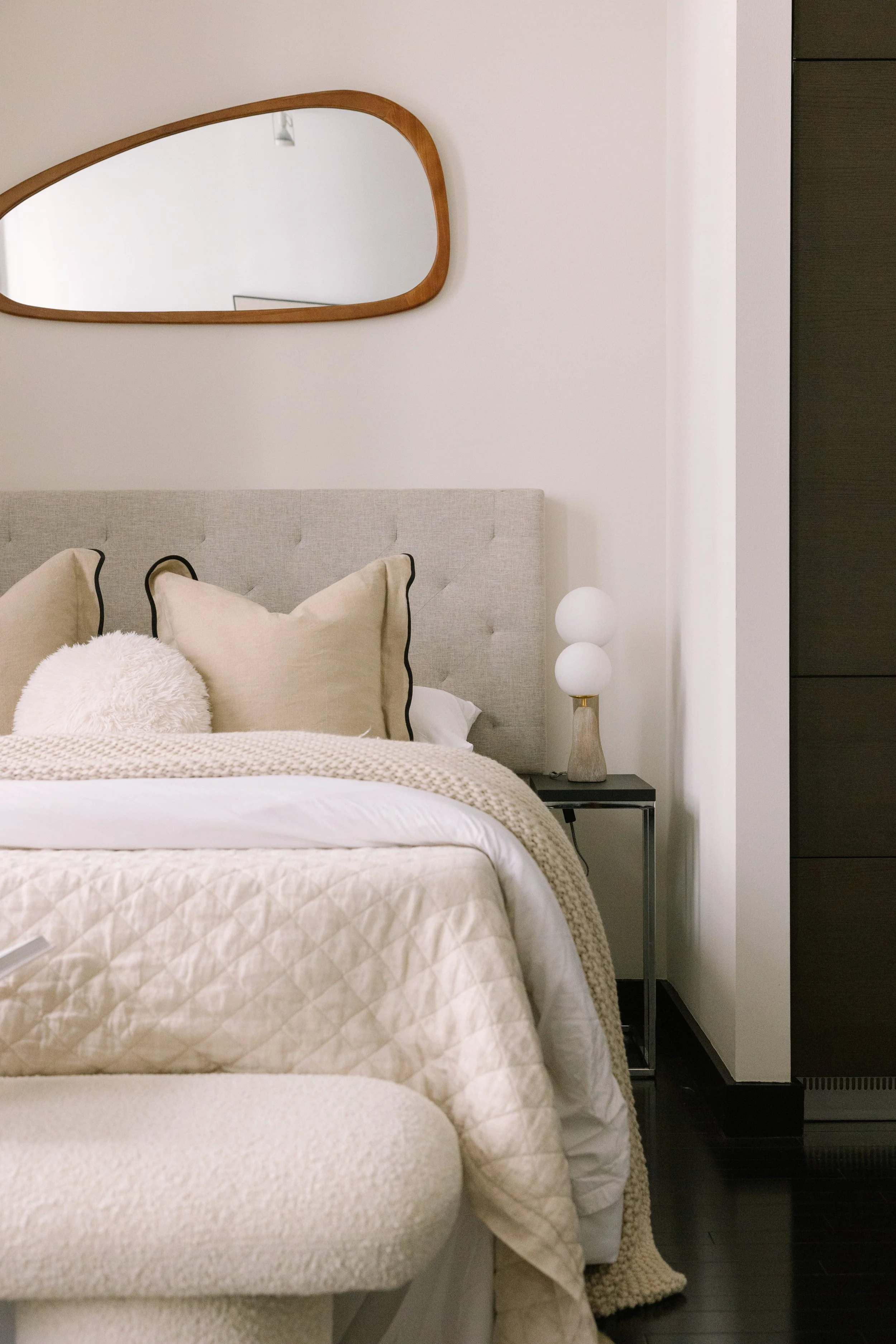 A cozy bedroom featuring a beige upholstered bed with throw pillows, a textured blanket, a modern bedside lamp with two white globes on a marble and black side table, a decorative wooden mirror on the wall, and dark hardwood flooring.