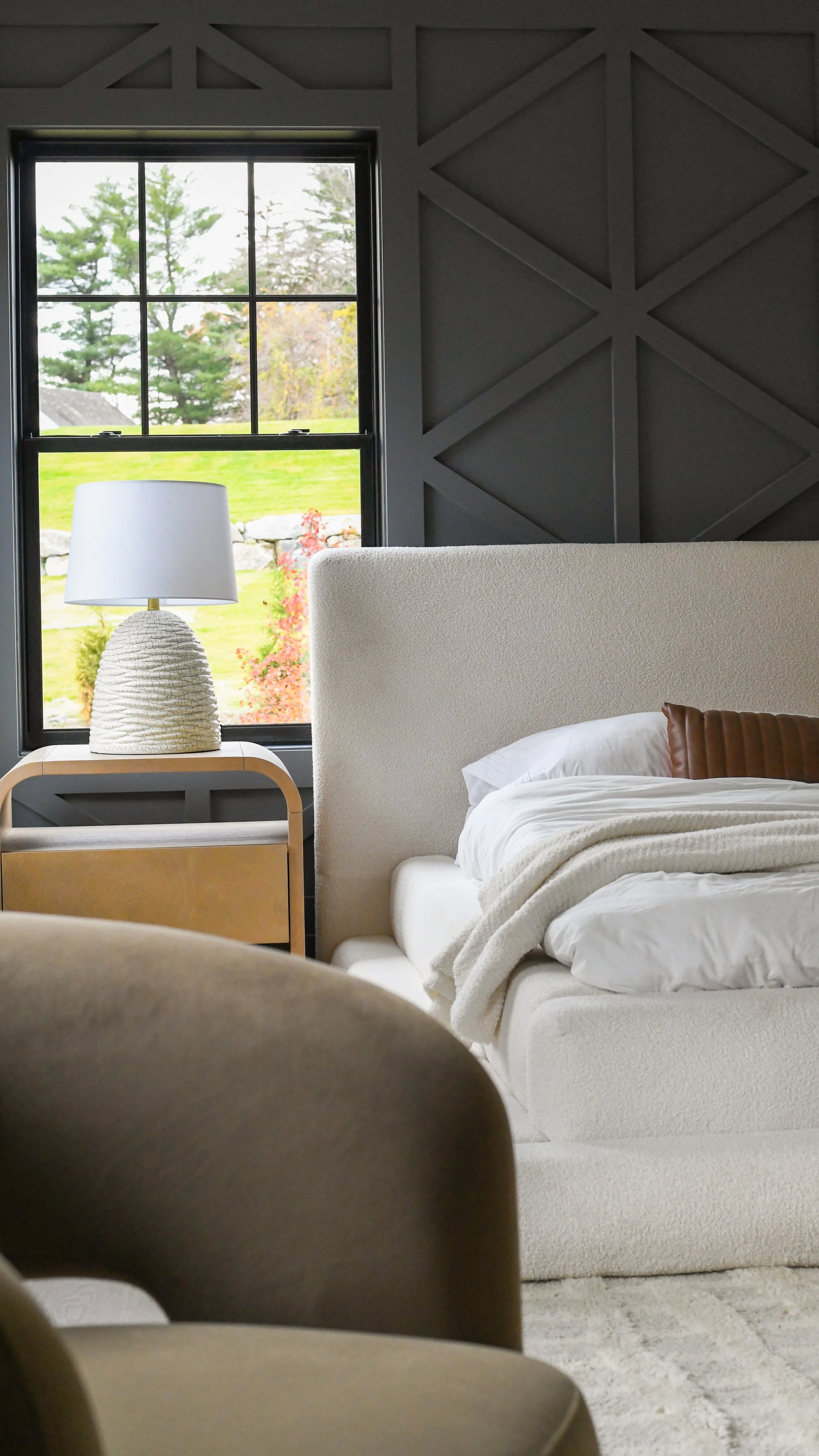 A cozy bedroom with a large window showing an outdoor scene with trees and grass. Inside, there is a white upholstered bed with a beige headboard, a folded blanket, and pillows. A wooden nightstand holds a textured lamp with a white shade.