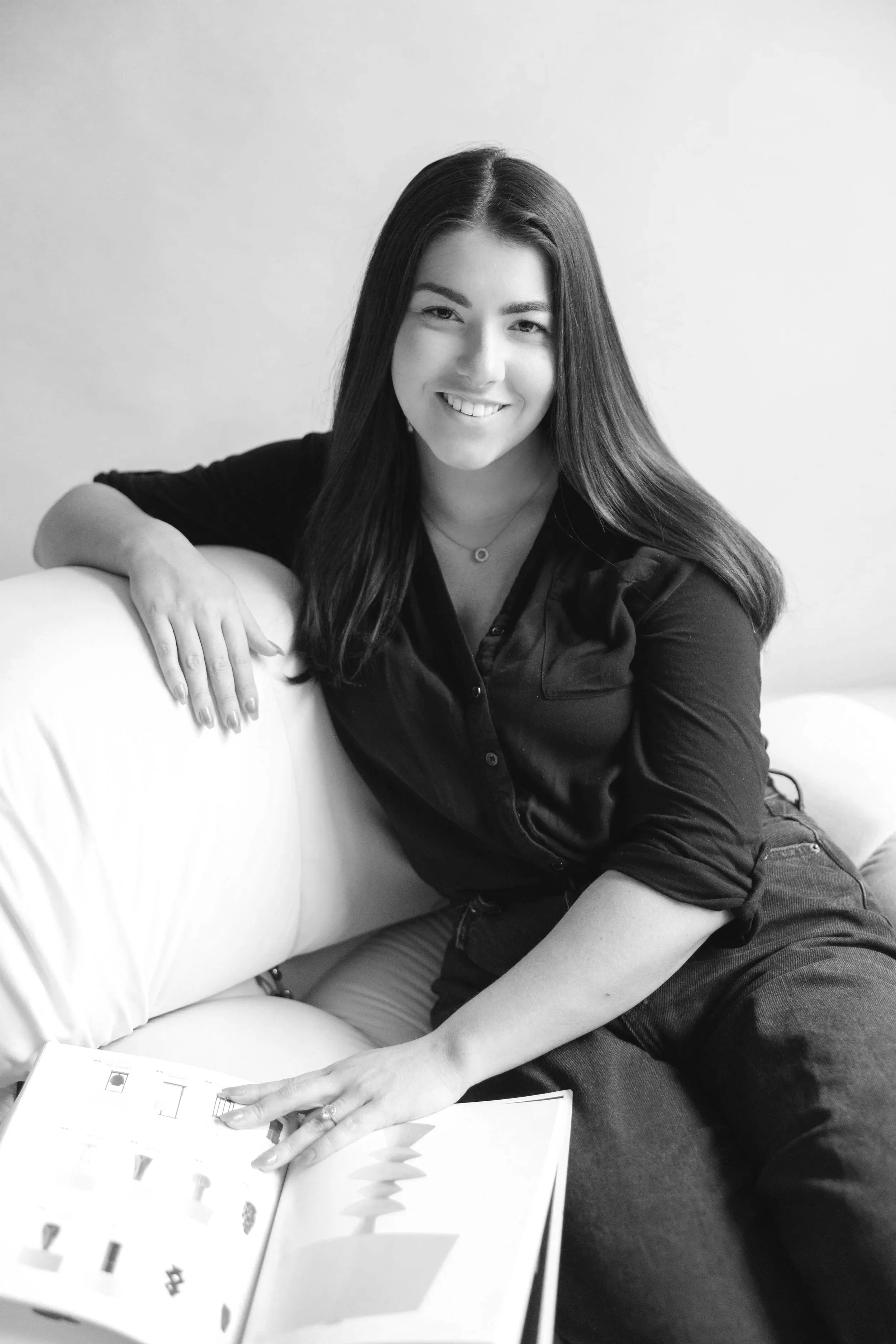 A young woman with long dark hair, smiling and sitting on a couch, pointing to a book open on her lap.