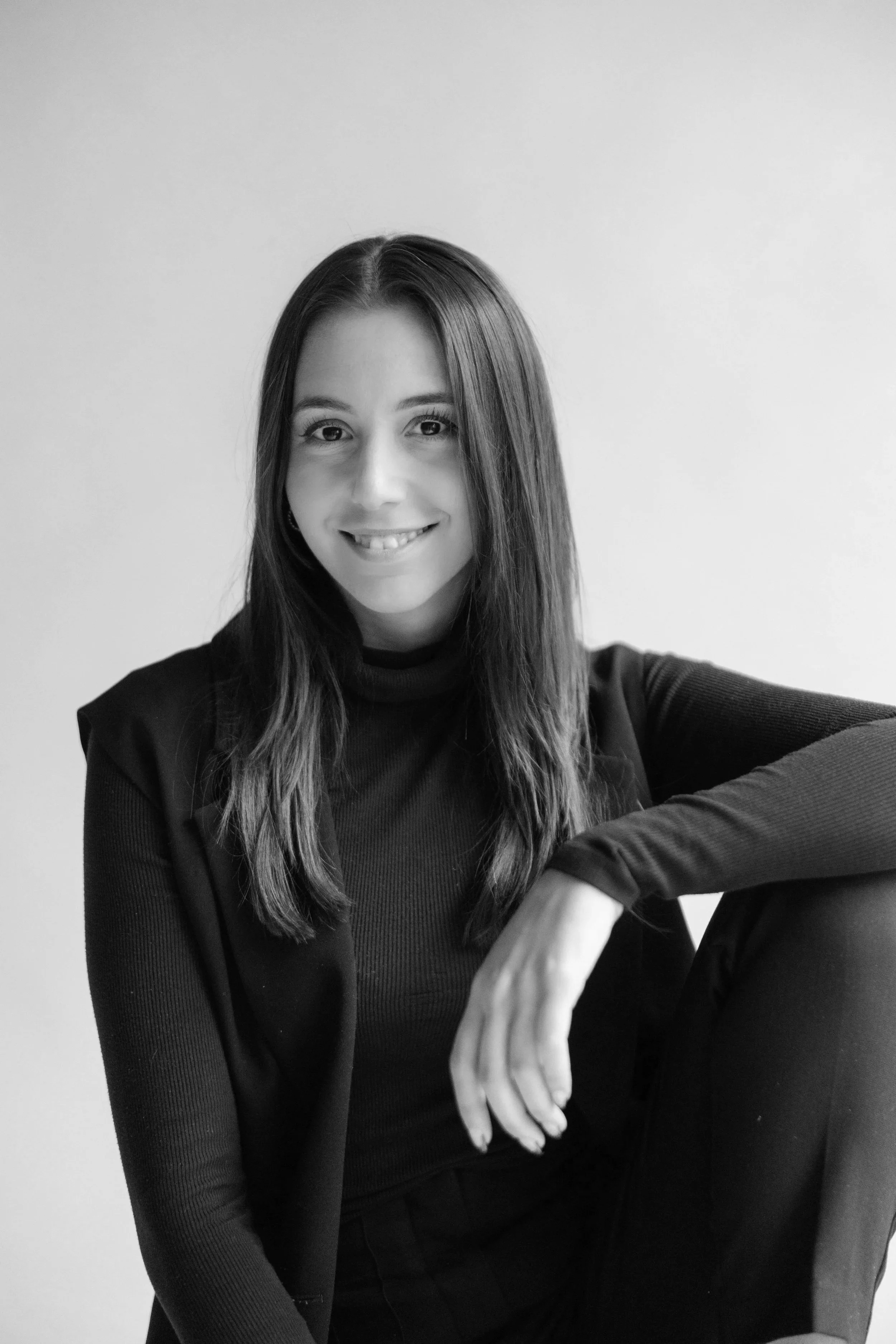 Black and white portrait of a smiling woman with long straight hair, wearing a dark turtleneck and jacket, sitting with one arm resting on her knee against a plain background.
