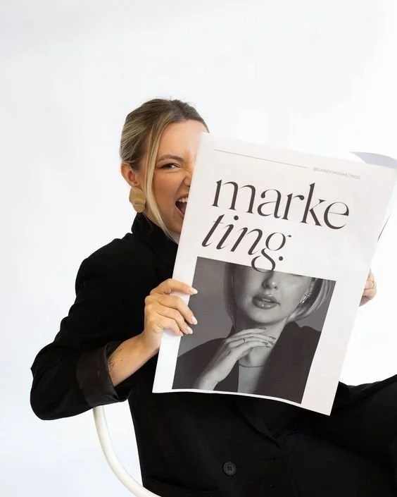 Woman smiling and playfully biting a large newspaper with the word 'marketing' on it, sitting against a white background.
