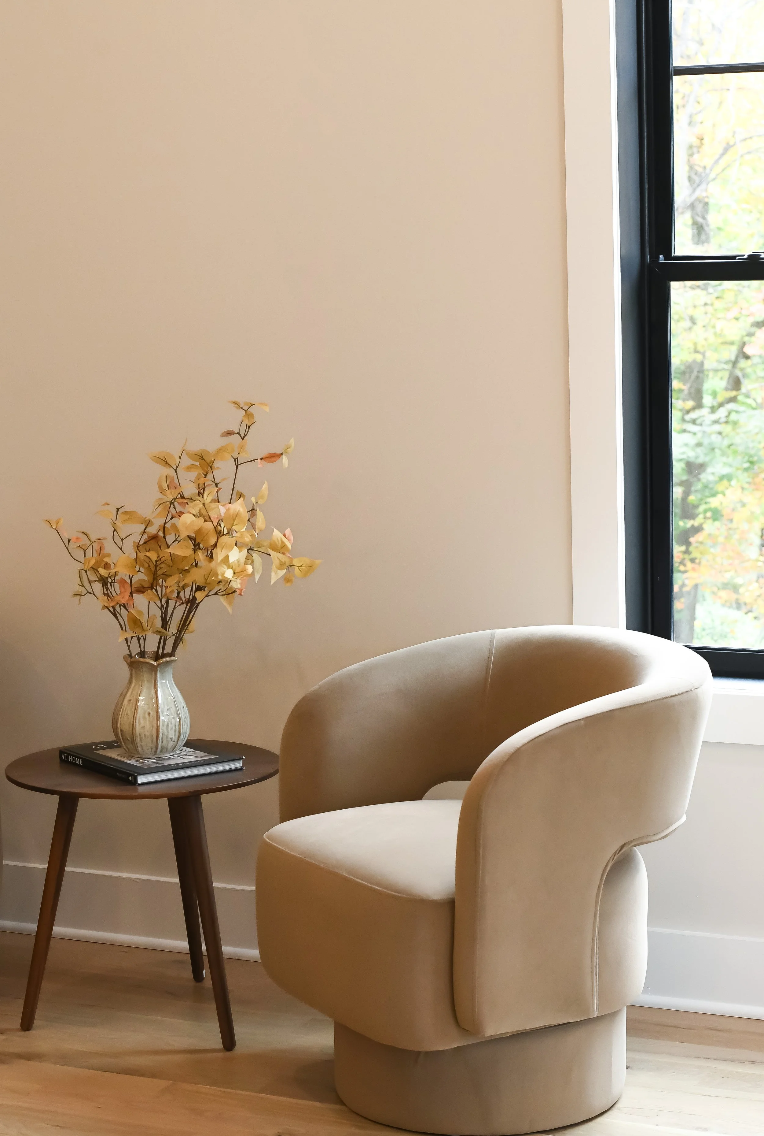A beige armchair next to a small round wooden side table with a vase of dried flowers and two books, near a window with black trim.