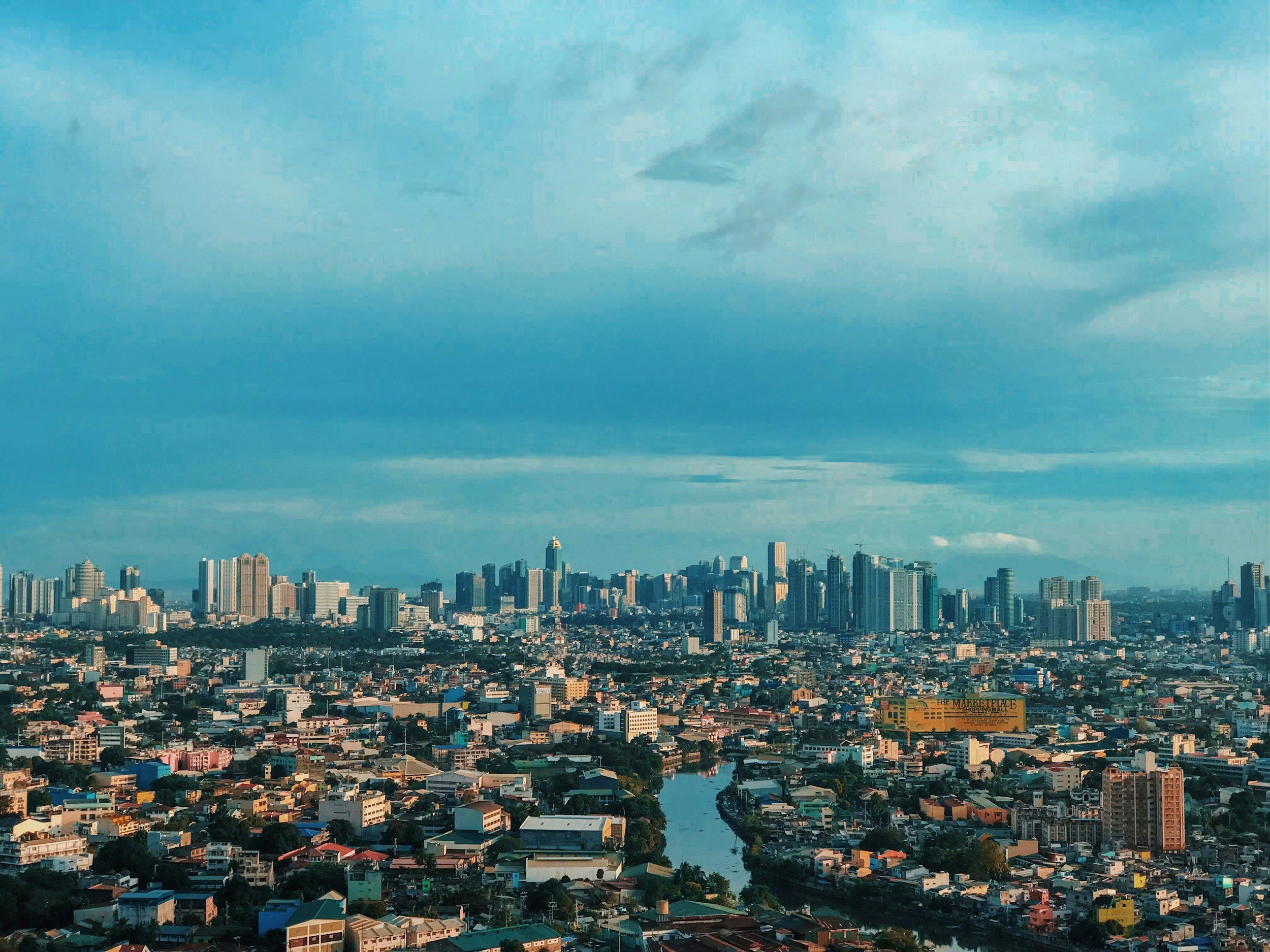 A panoramic view of a city skyline under a blue sky with clouds, featuring high-rise buildings, residential areas, and a river flowing through the city.