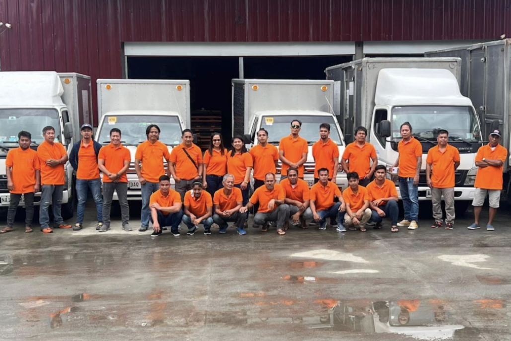 Group of people in orange shirts standing and kneeling in front of delivery trucks outside warehouse.