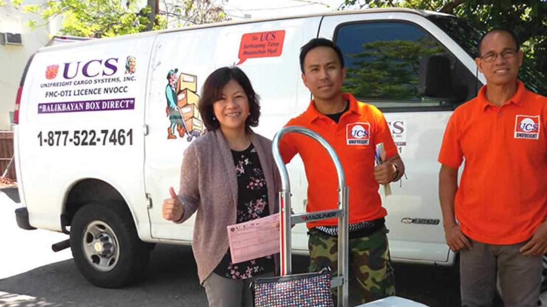 Three people standing in front of a UCS delivery van, two men in orange shirts and a woman holding a document, with a cart in front of them, outdoors with trees and a fence.