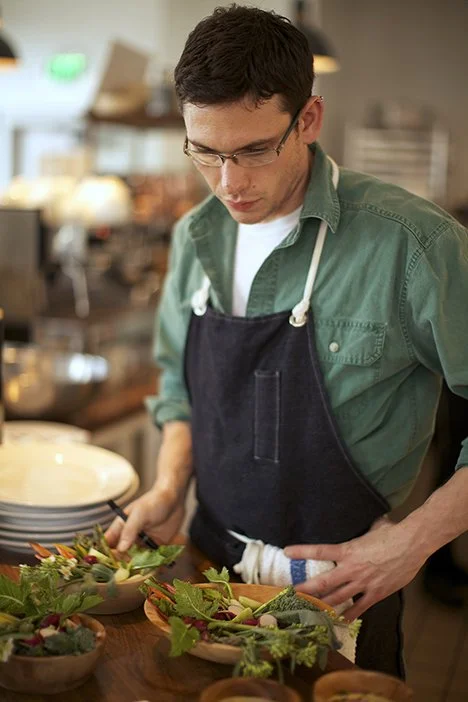 A man wearing glasses, a green shirt, and a dark apron is preparing or plating food at a restaurant or café.