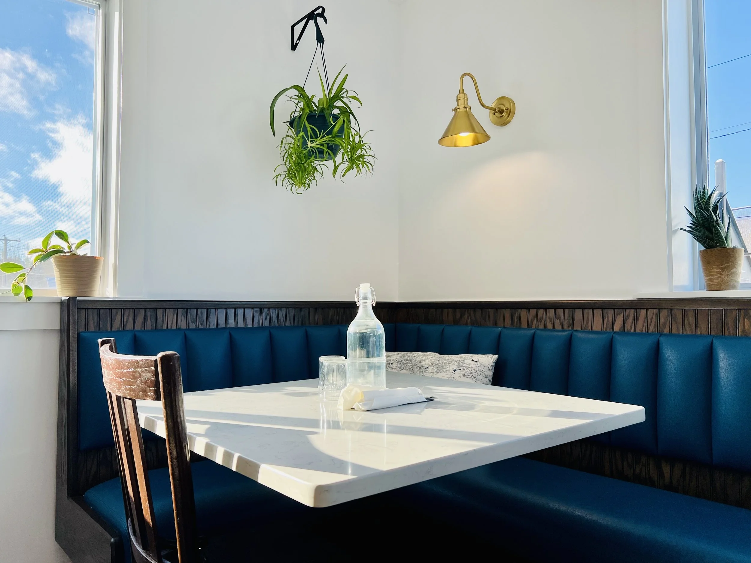 Empty restaurant booth with blue upholstered seating, white marble table, green plants on windowsill, hanging plant, and a wall-mounted gold lamp, with sunlight streaming through windows.
