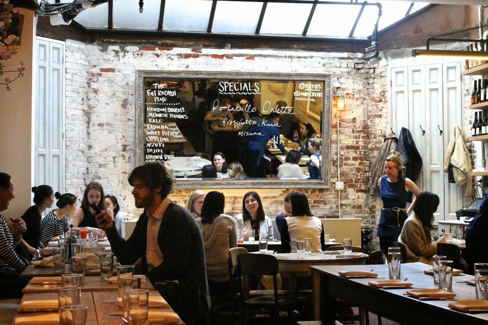 Bustling restaurant interior with exposed brick walls, a large mirror, and patrons seated at wooden tables set with glasses and napkins, some team members are standing and interacting with customers.