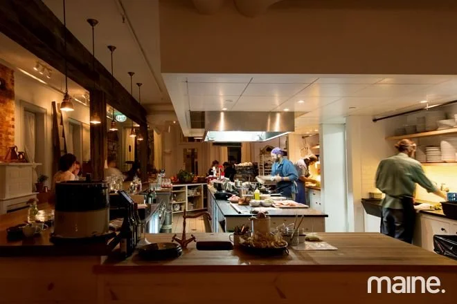 A busy restaurant kitchen with chefs preparing food, open layout, wooden counter in the foreground, and warm lighting.