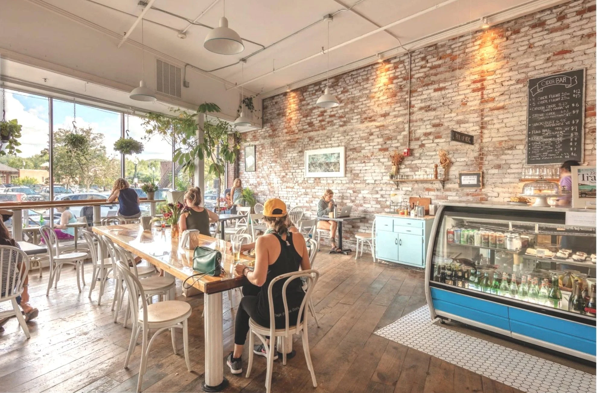 Interior of a bright coffee shop with exposed brick walls, large front windows, hanging plants, and white furniture. Customers are sitting and working or socializing.