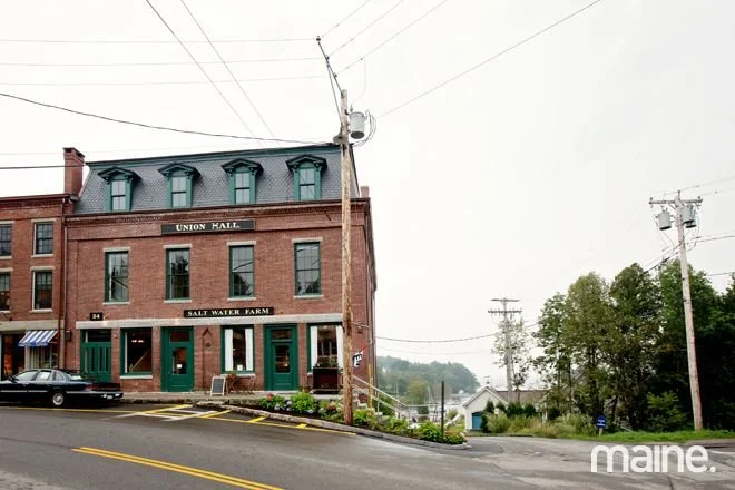 A three-story brick building labeled 'Union Hall' and 'Salt Water Farm' with green doors and windows, located on a sloped street with parked cars, utility poles, and trees in the background.