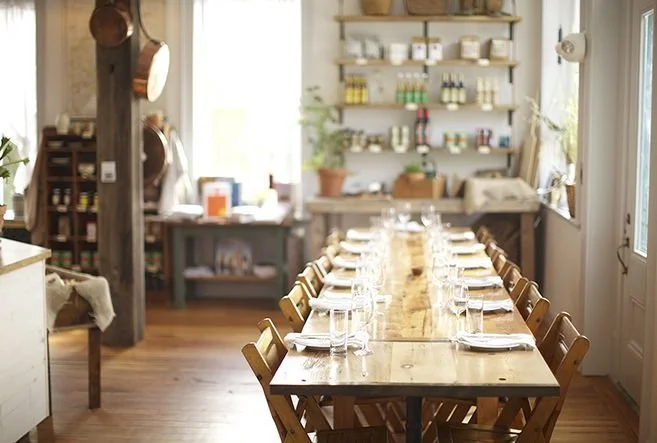 A dining room with a long wooden table set with plates and glasses, surrounded by wooden chairs, and decorated with shelves holding jars and bottles, with sunlight coming through the windows.