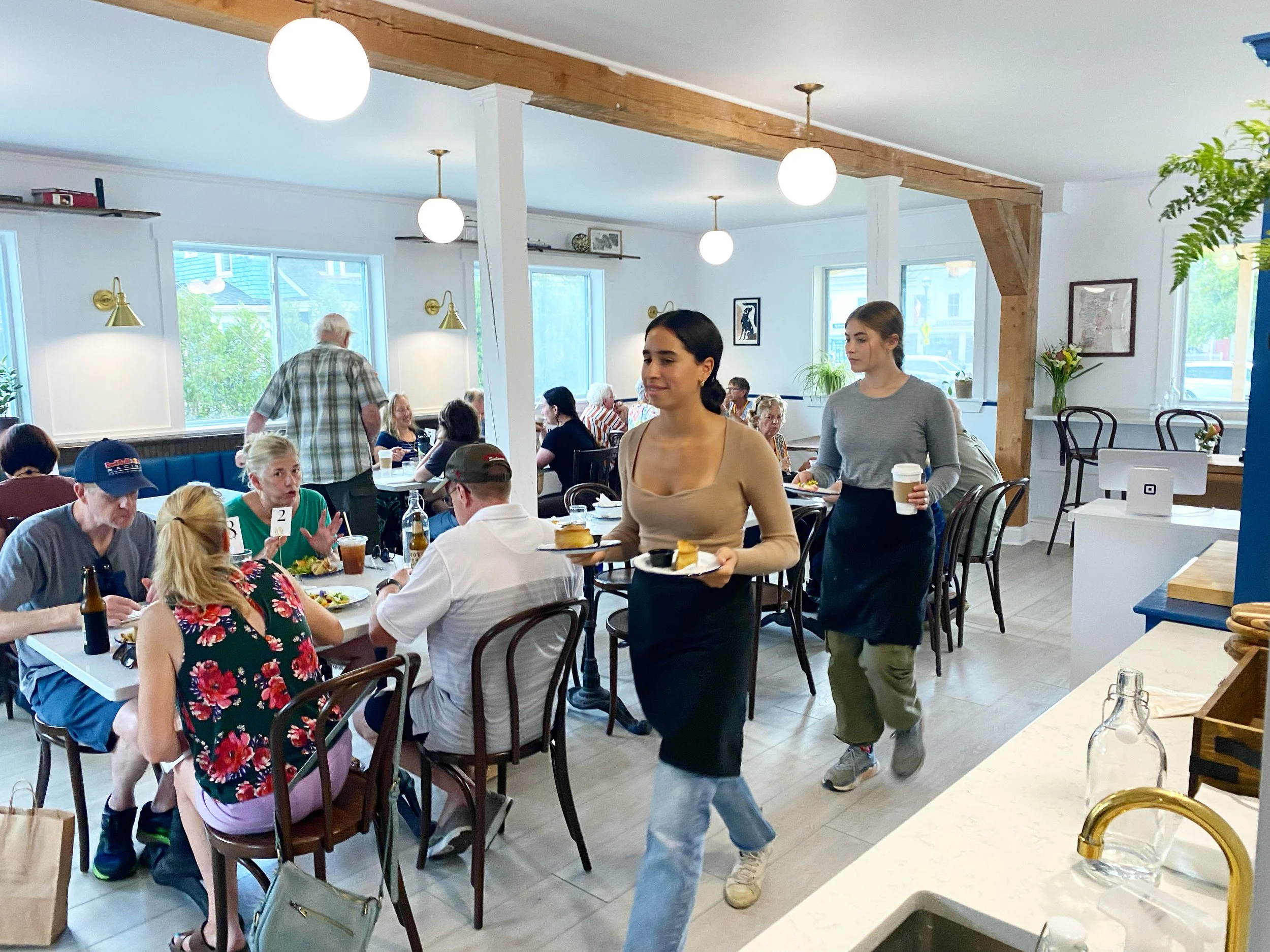 People dining in a bright restaurant, seated at tables with food and drinks, while waitresses serve patrons.