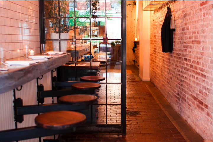 Interior view of a cozy restaurant with a rustic and industrial design, featuring a brick wall, wooden bar stools, a metal and glass partition, and a dining area with table settings visible in the background.