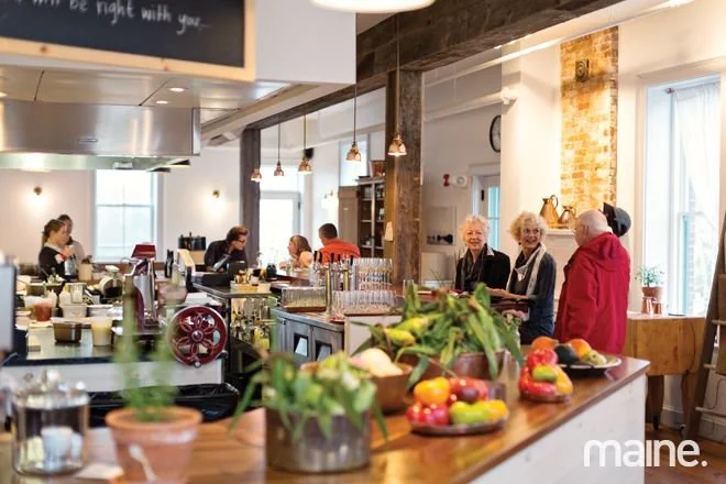 People socializing and dining in a cozy cafe with a counter filled with fresh fruits, plants, and kitchenware, illuminated by hanging pendant lights and natural light from large windows.
