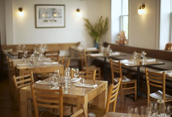 Unoccupied restaurant dining area with wooden tables, chairs, and neatly arranged glassware and silverware.