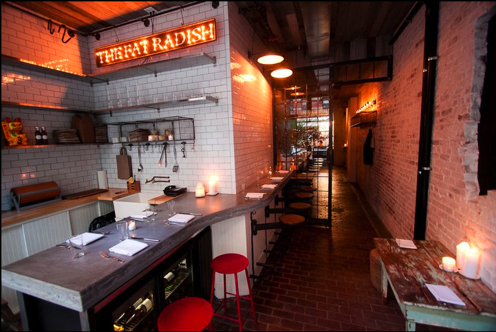 Interior of a restaurant with a bar counter, stools, shelves with glasses and plates, a neon sign that reads 'THE FAT RADISH', candles on the counter and table, brick and tiled walls, and pendant lighting.