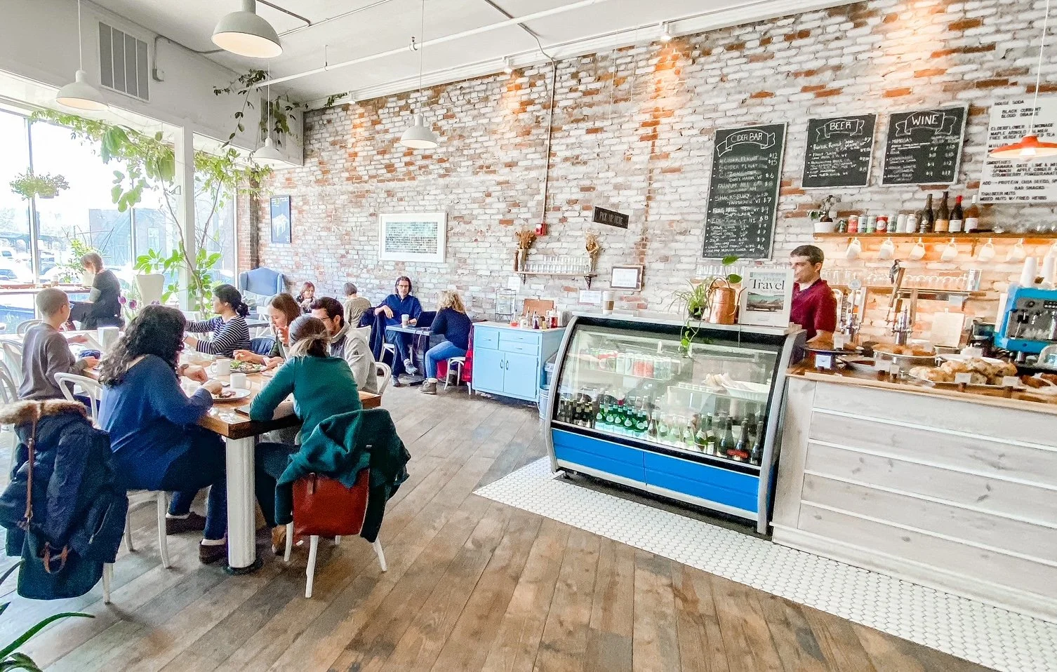 Inside a cozy cafe with exposed brick walls, several customers seated at tables, large front windows allowing natural light, a refrigerated display case with drinks, a barista behind the counter, and a chalkboard menu on the wall listing drinks and p