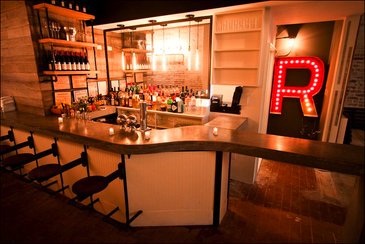 A modern bar with a curved marble counter, shelves with bottles of alcohol, and a large illuminated red letter 'R' in a corner.