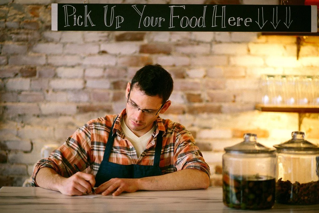 A man wearing glasses and a plaid shirt with an apron, writing or signing on a piece of paper at a counter. There are glass jars with food or drinks on the right side. A sign above reads "Pick Up Your Food Here!" with arrows pointing down.