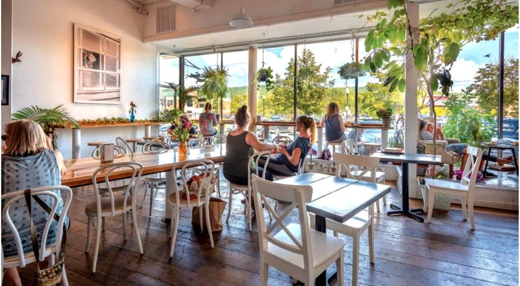 People inside a brightly lit cafe with large windows and outdoor seating, decorated with plants, sunlight, and casual seating arrangements.