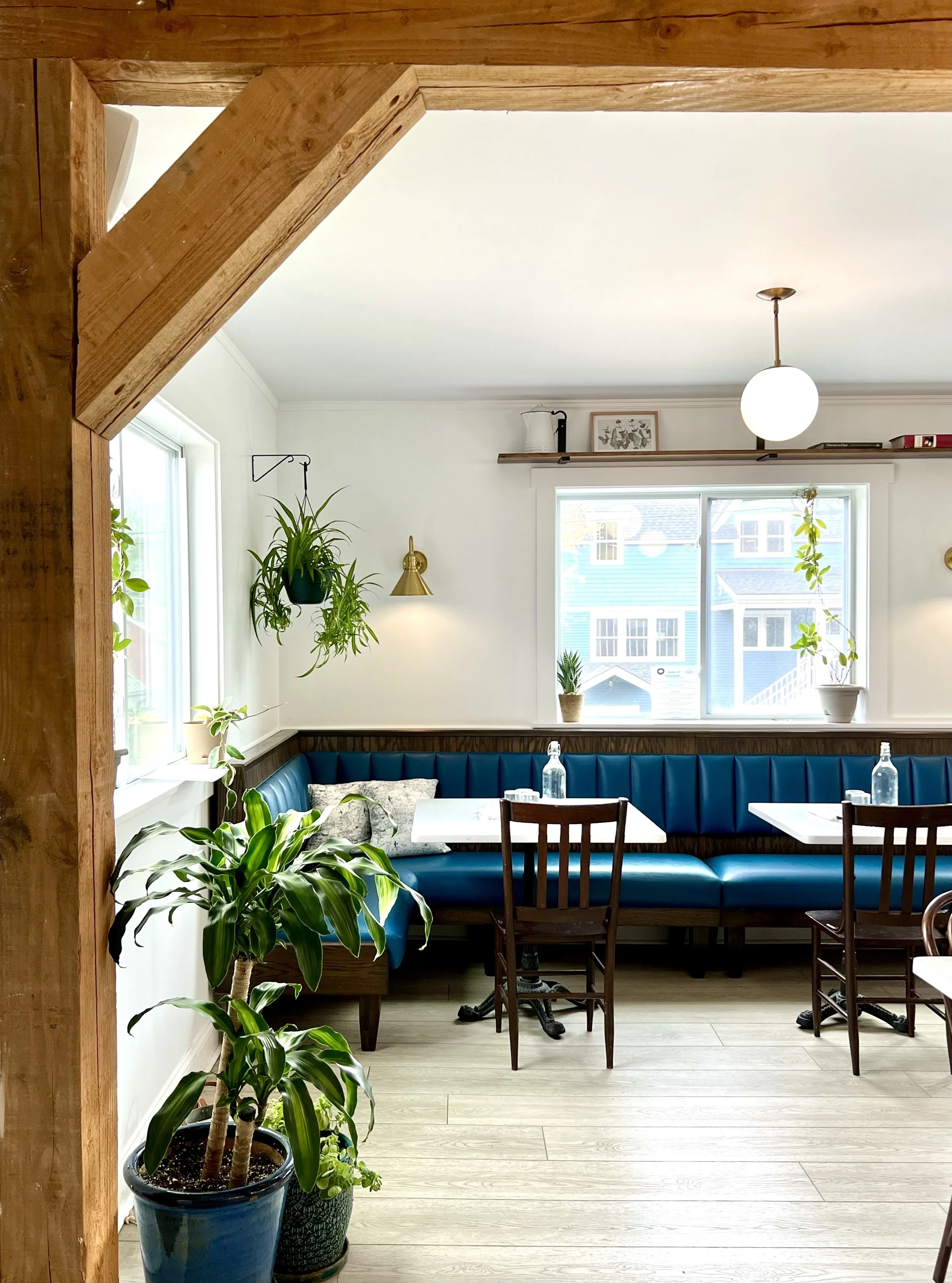 Cozy dining area with white walls, a large window, green plants, a blue upholstered bench, wooden chairs, and modern decor.