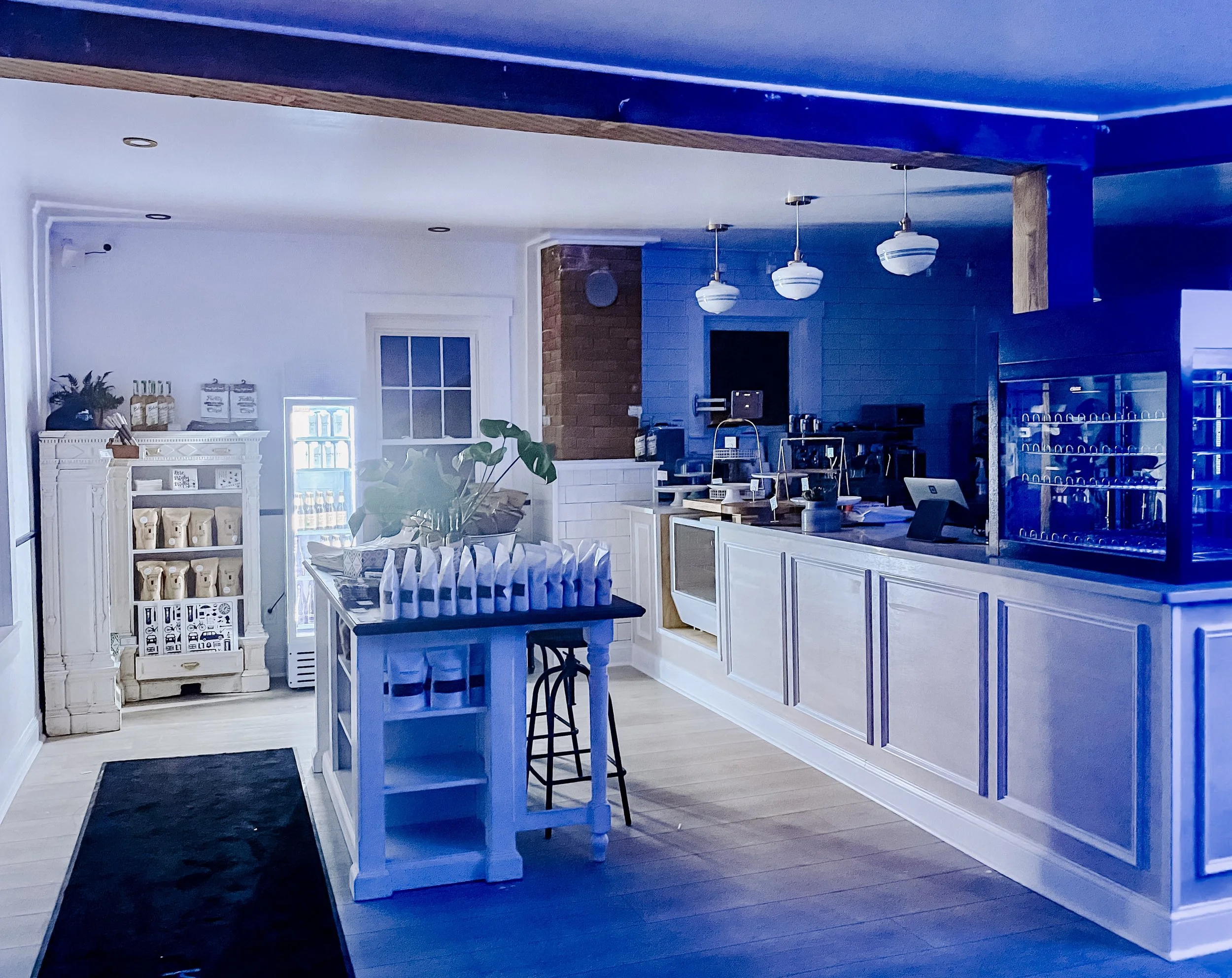 Interior of a modern cozy cafe or restaurant with a white wooden cabinet, a dining table with a plant, hanging pendant lights, and a counter with a point of sale system. The space features white walls, a blue ceiling, and wood accents.