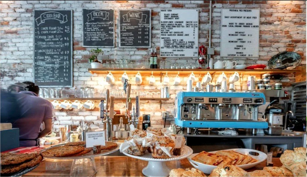 A cozy coffee shop with a brick wall, black chalkboard menus displaying beer, wine, and food options, an espresso machine, and various baked goods on a wooden counter.