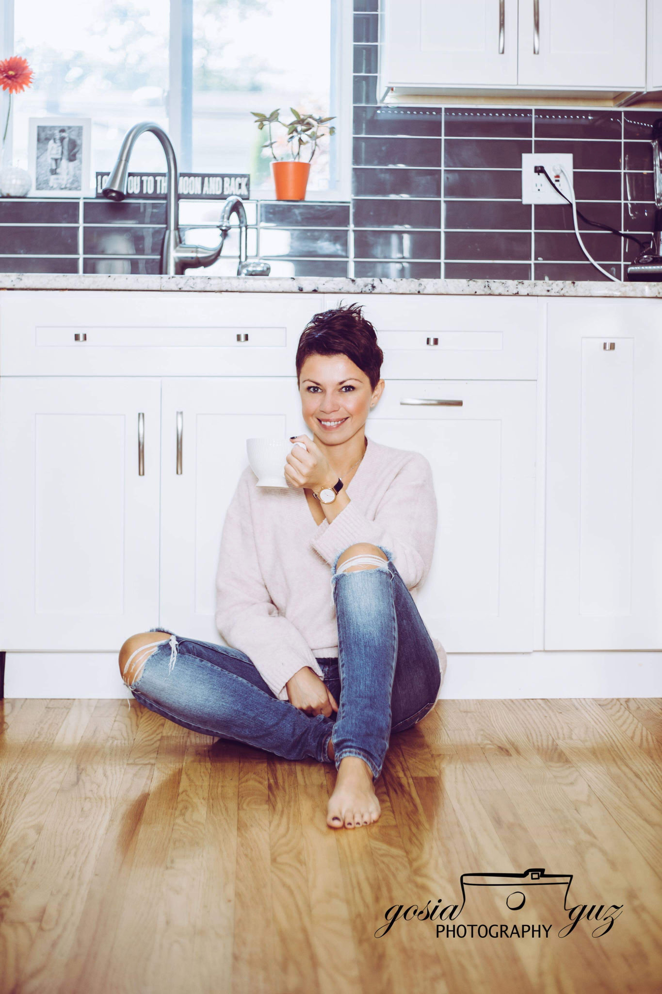 Woman sitting on a wooden floor in a kitchen, holding a white mug, smiling, wearing a pink sweater and ripped jeans.