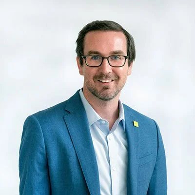 Portrait of a man wearing glasses, a blue suit, and a white shirt, smiling against a plain light background.
