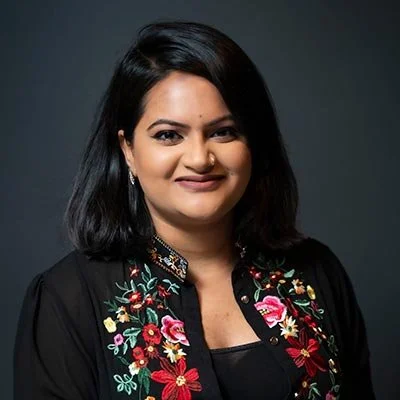 Portrait of a woman with shoulder-length black hair, wearing a black top with floral embroidery, smiling against a dark background.