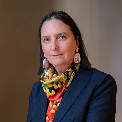 Portrait of a woman with dark hair, wearing a navy blazer, colorful scarf, and large earrings.