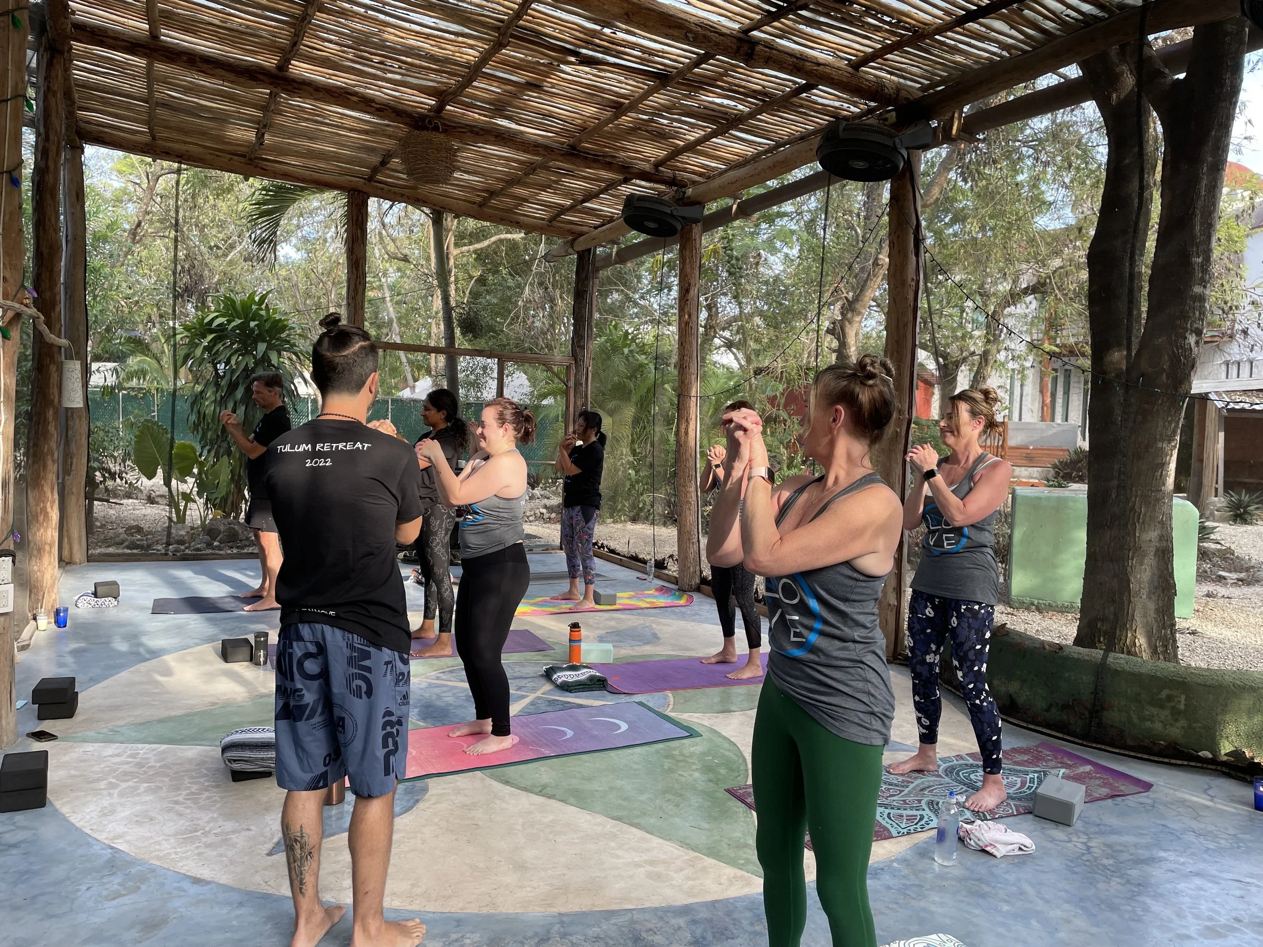A group of people participating in a yoga class outdoors, standing on yoga mats in a shaded, open-air structure with trees and greenery outside.