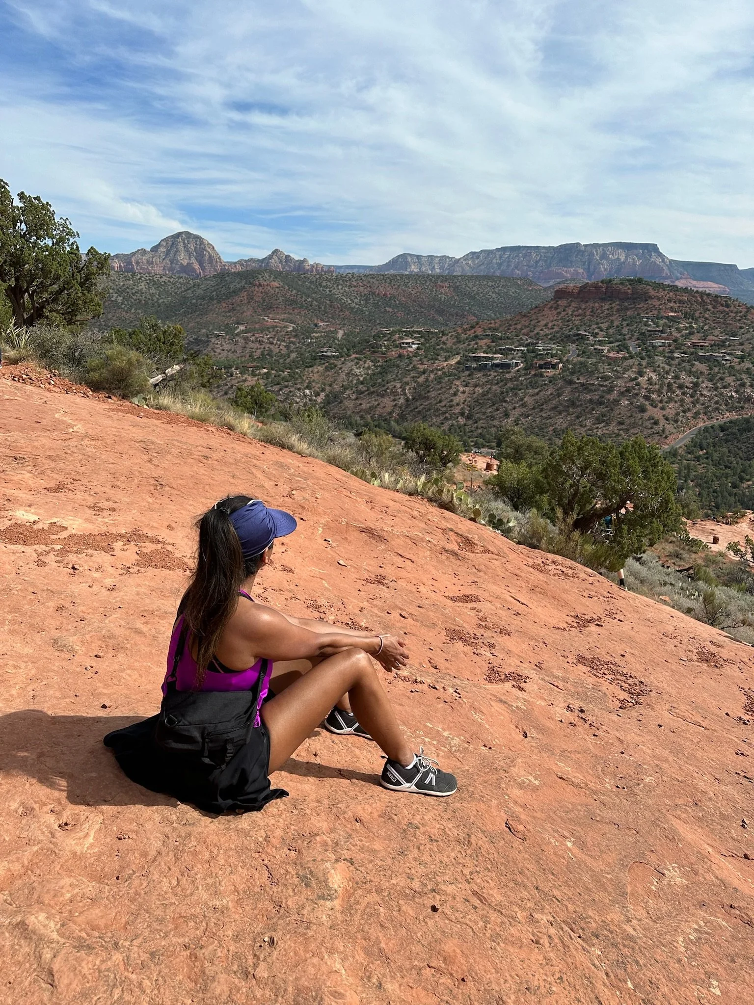 A woman sitting on a red rock slope in a desert landscape with mountains and blue sky in the background.