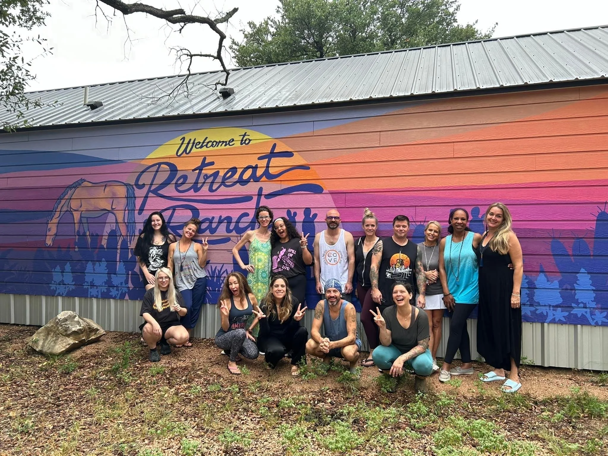 Group of people smiling and posing in front of a colorful mural that reads 'Welcome to Retreat Ranch.' The group includes men and women dressed in casual and summer clothing, standing on dirt ground with some greenery and rocks.