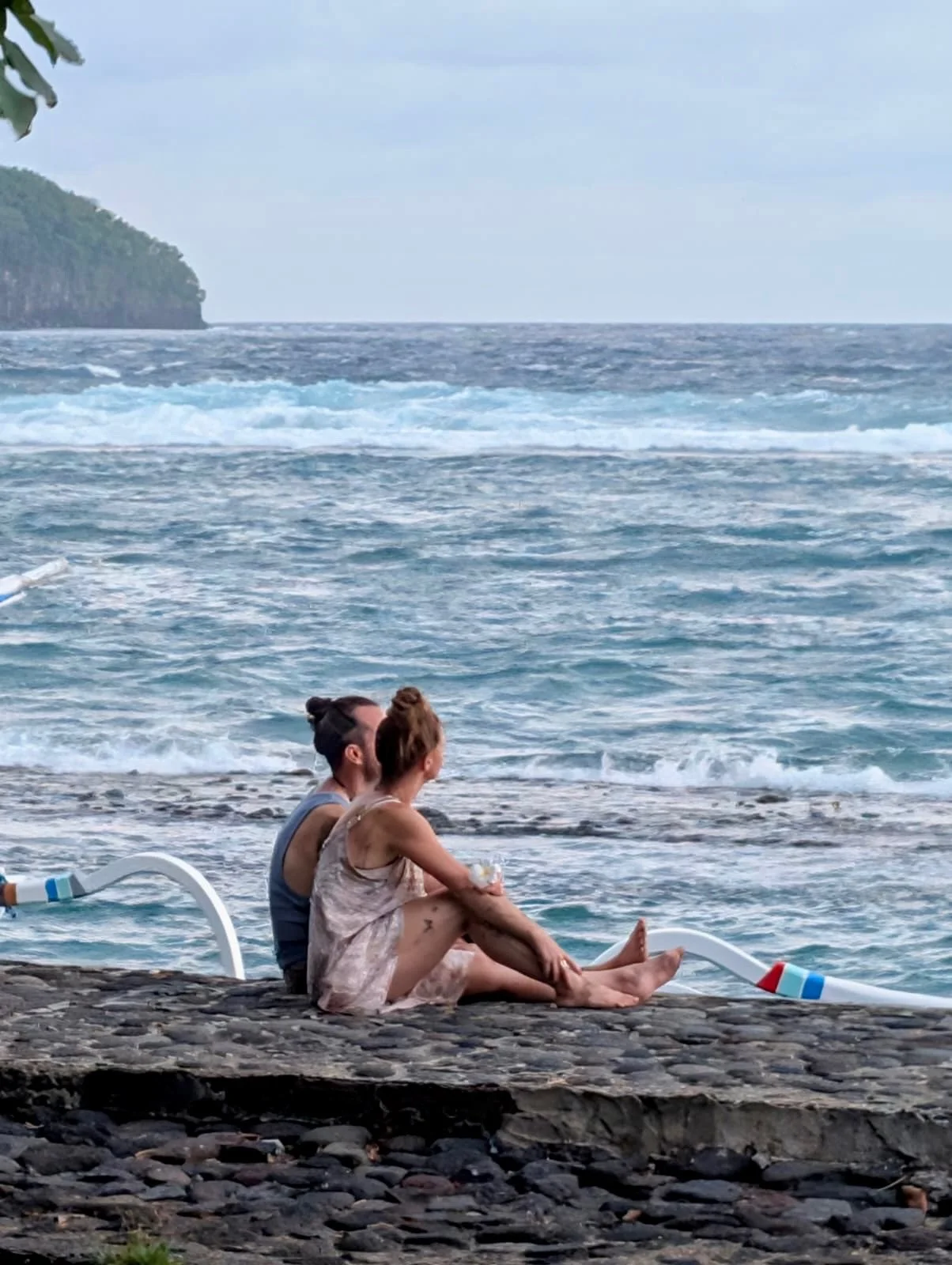 A couple sitting on a rocky beach near the ocean, with waves in the background and a distant green headland.