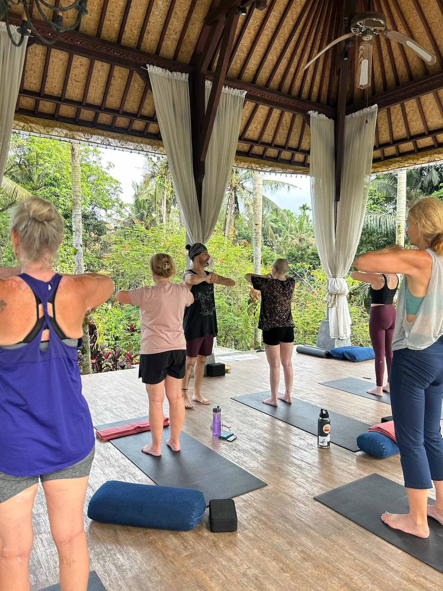 Group of people practicing yoga on mats in an open-air pavilion with wooden ceiling and white curtains, surrounded by tropical greenery and palm trees.
