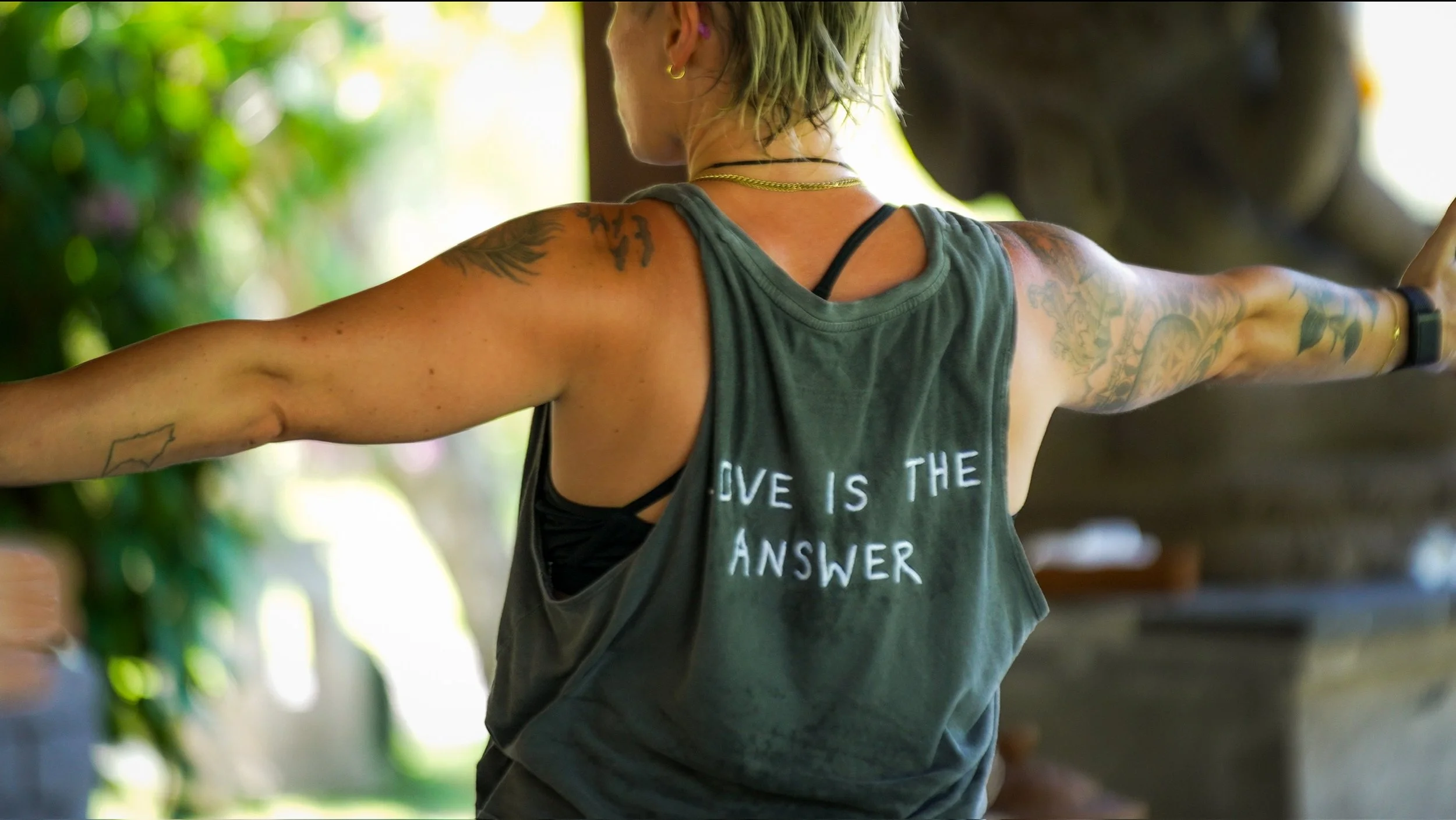 Back view of a woman with short, dyed green hair and tattoos, wearing a sleeveless gray top with the slogan 'LOVE IS THE ANSWER'. She has her arms outstretched and appears to be outdoors in a sunny, green environment.
